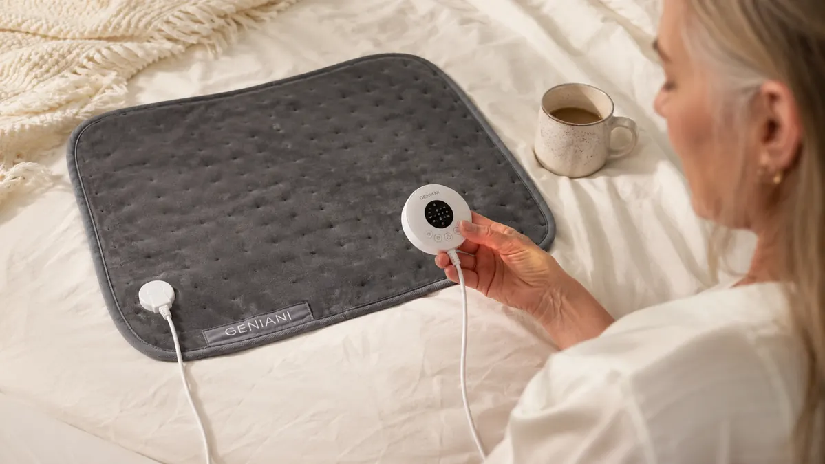 A hand turning a simple dial control on a gray electric heating pad resting on a bedspread beside a cup of coffee