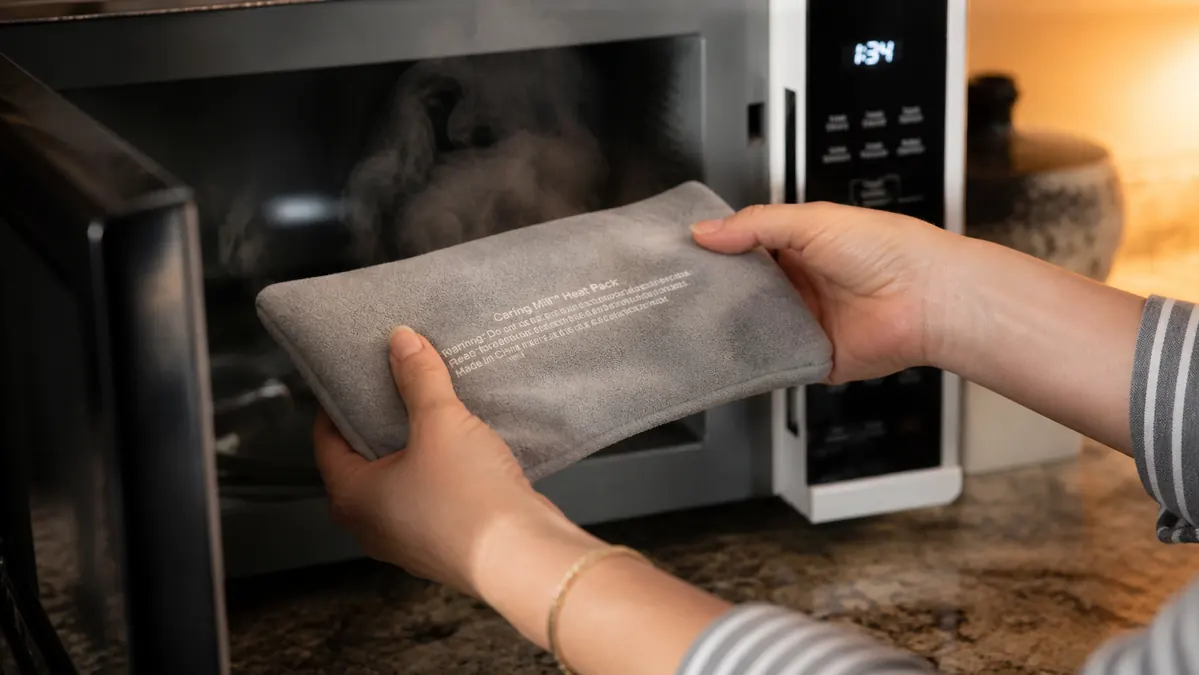 Hands taking a gray gel heat pack out of a microwave, steam rising gently, in a kitchen with warm light