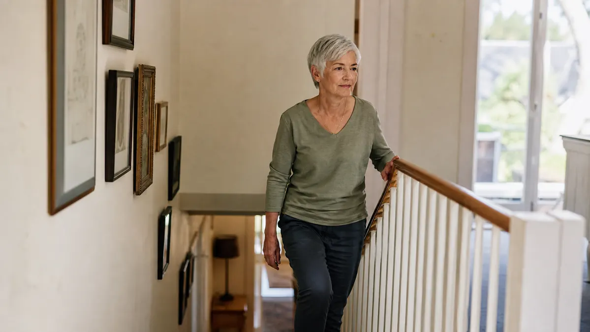 An older person walking up a short flight of stairs at home, one hand on the railing, wearing a fitted knee sleeve under their trousers